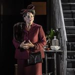 Older woman, The Princess (Anne Mason) with sour expression on face in red dress suit and hat, hands clasped before her, standing next to a staircase