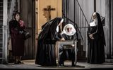 A nun and a woman in red dress suit watch as a nun sits at a desk, her hand held to the desk by another nun.