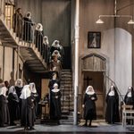 A staircase and hallway of sparse nunnery, lined with nuns, looking down at a nun singing