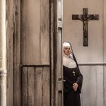 The Abbess (Sioned Gwen Davies) lurks in a doorway below a cross