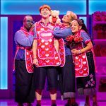 Robbie (Blake Patrick Anderson) in shop bib with three store women cuddling up to him with roses, and teddy bears