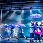 Three shop women huddle under purple umbrellas as others dance with purple umbrellas in the background