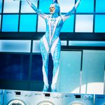 Jem (River Medway) in blue and white morph suit standing on a row of washing machines, arms thrown up