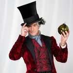 Drosselmeyer (James Streeter) tipping his top hat, holding a large dark copper bauble in his other hand, wearing red paisley waistcoat and jacket