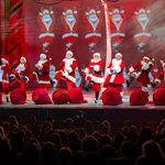 Ten Santa's in a line kicking at large red sacks at the edge of the stage, shadowed audience in the foreground