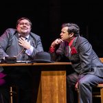 Del Boy (Sam Lupton) in suit, leaning on cluttered table listening to Dating Agent (Richard J Hunt) intently