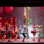 Humans dressed as elves spinning below aerial cirque star, fireworks on screen behind them