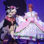 Dame Trot (Matt Rixon) in large white dress with pink flowers, with pantomime cow with pink bow and collar next to her