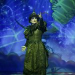 Mrs Blunderbore (Anne Smith) wearing black dress and hat, washed in green light, pointing menacingly forward with crescent moon face backdrop behind