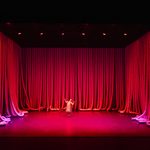 Red lit stage dressed with industrial air-tubing hanging down on all sides, a dancer alone in the middle