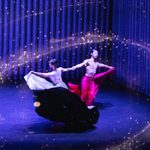 Male Asian dancer in long red skirt clasps wrists with woman in long silver sparkly skirt, counter-balancing as they spin surrounded by gold sparkles swirling in the air around them