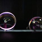 Two Asian women in long skirts and mismatched socks dance as projections of large bubbles hover before them