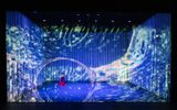 Male Asian dancer in long red skirt thrusts hand towards water illusion making a swirling circular hole in the illusion to the left