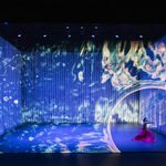 Male Asian dancer in long red skirt thrusts hand towards water illusion making a swirling circular hole in the illusion to the right