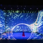 Male Asian dancer in long red skirt thrusts hand towards water illusion making a swirling circular hole in the illusion in the centre of the stage