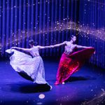 Male Asian dancer in long red skirt clasps wrists with woman in long silver sparkly skirt, counter-balancing as they spin surrounded by gold sparkles swirling in the air around them