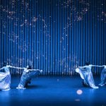 Two pairs of dancers in long sparkly silver skirts counter-balance spin on a dim stage with gold sparkles in the air