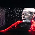 Projection of person with short hair, eyeliner and red lips in red top, projected onto bumpy stage background with sparkles falling around