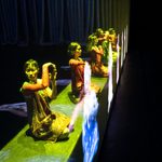 Six Asian women kneel making right angles with their hands in front of them as holograms of figures hover before them