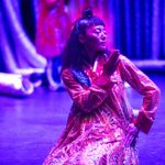 Asian woman kneels, left hand on hip, right hand gesturing away from the opposite shoulder