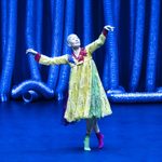 Shaved-headed Asian woman in yellow dress, one green sock and one pink at the top of a curtsey-like action as she dances
