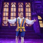 Silly Simon (Kev Orkian) in blue and orange striped waistcoat spreads his arms wide with big smile in front of big house