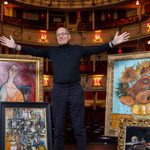 Peter James, arms spread wide, in theatre, surrounded by paintings