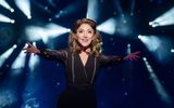 The Narrator (Christina Bianco) in black outfit, reaches out towards the camera, lights shining behind her.