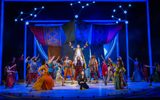 Joseph (Adam Filipe) in the technicolor coat, sitting on the shoulders on one of his brothers as the others and ensemble point up at him, the Narrator (Christina Bianco) kneels in front.
