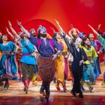 Company and Narrator (Christina Bianco) one hand on hip, the other raised in the air, in a desert area with orange sky backdrop