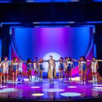 Company in cheer outfits, one arm extended to point to Joseph standing centre stage in his technicolour coat, arms spread wide. Spotlights dot the purple lit stage.