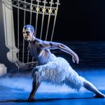 The White Swan, a bare chested man in white feathered trousers, lunges to his right on a foggy night with pillars and gates in the background