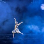 The White Swan, a bare chested man in white feathered trousers, leaps over wavy water on a misty full moon night