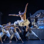 Back to camera, the White Swan leaps, one leg and arm forward, the other arm raised high. The other swans, all bare chested men wearing white feathered trousers, watch his or climb on the royal bed