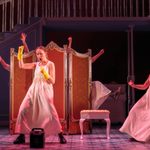 A young white woman in white dress and marigolds cheers next to a karaoke speaker, a similar woman pumps her fists to the side, behind a changing screen two people throw their legs and arms out.