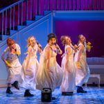 Five Regency women in white dresses and Docs, singing into hand mics with a karaoke machine speaker in front of their v formation