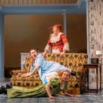 A Regency red-dressed ginger white woman stands behind a sofa, a blonde white woman in blue regency dress reclines on the sofa, a blonde white woman in green regency dress falls the other way on the floor by the sofa, all look startled.