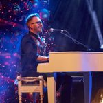 White man in gloasses and plaid waistcoat plays white piano, smiling, large Christmas tree behind him