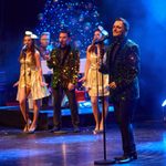 White man in glittery green jacket with standing microphone, other singers behind him