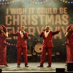 Two white women in long red dresses with hip slits bracketed by two white men in red suits, singing arms outstretched as snow falls and the screen behind them reads I wish it could be Christmas everyday