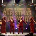 Two white women in long red dresses with hip slits bracketed by two white men in red suits, singing arms outstretched as snow falls and the screen behind them reads I wish it could be Christmas everyday