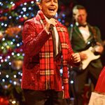 White man in plaid red scarf and red sparkly jacket singing into hand mic