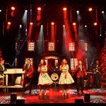 White women in pale green tea dresses bracketed by white men in red sparkly jackets singing into hand mics with band (piano, guitars, drums) and large Christmas trees and house facade behind them.