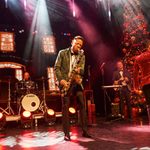 Black man in dark green jacket enthusiastically plays the sax as couples dance