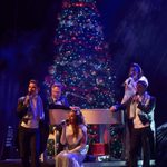 Five people around a piano in formal wear, a large Christmas tree behind them