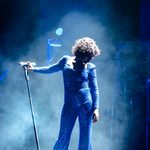 Whitney Houston impersonator looks down to the side as she thrust the mic stand to the side in blue haze