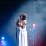 Whitney Houston impersonator in long white dress holding standing mic, smiling, side profile with smoky stage lights behind