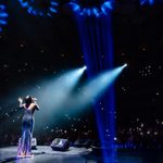 Rear view of woman in floor-length shoulderless dress, singing into hand mic before dark auditorium filled with phone lights