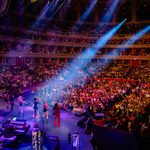 Aerial view from back of stage of six singers and band performing to packed theatre