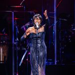 Whitney Houston impersonator in black klong shoulderless dress singing into standing mic